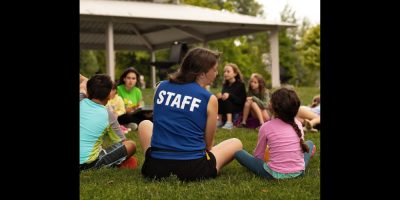 A camp staff member sitting in a circle with young children on grass.