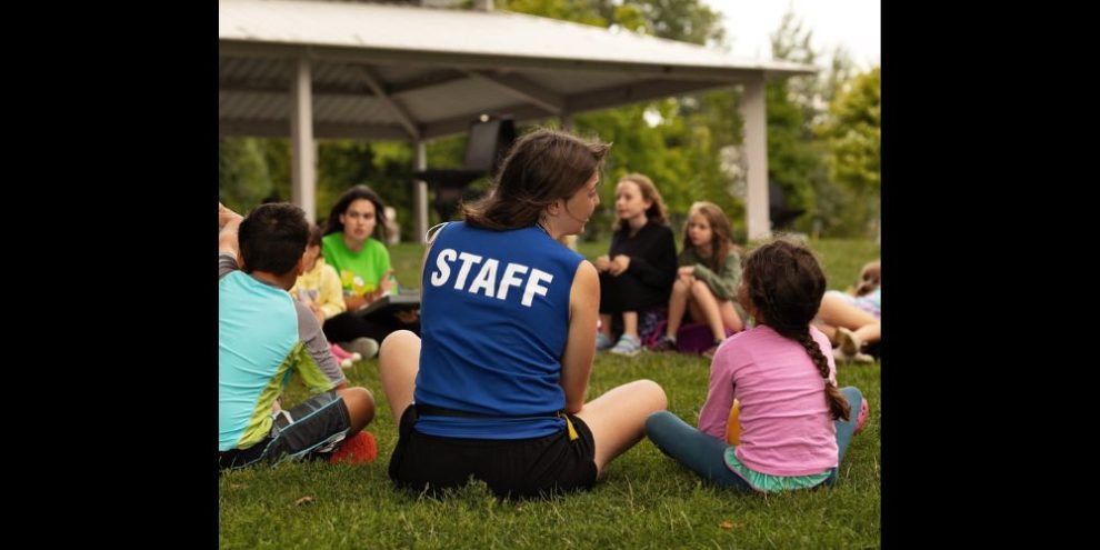 A camp staff member sitting in a circle with young children on grass.