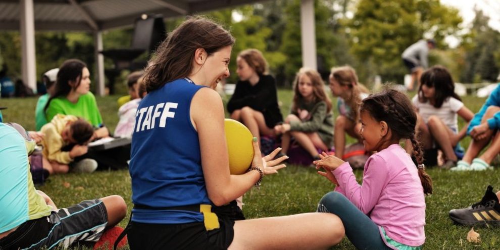 Summer camp staff member from the City of Barrie holds a ball while a child watches during an outdoor activity.