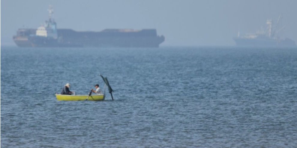 Two men in a small boat with cargo ships anchored in the Strait of Hormuz near Bandar Abbas, Iran