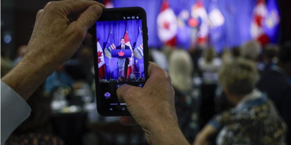 A person uses a smartphone to photograph a political speaker during a public event, illustrating how Canadians consume news and social media content on mobile devices.