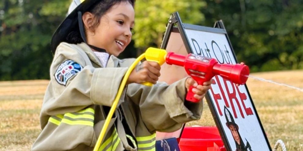 Young girl wearing a firefighter jacket and helmet sprays water from a toy fire hose at a Barrie Fire Hot Summer Nights event.
