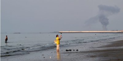 Smoke rises over the Strait of Hormuz near Bandar Abbas, Iran, as a woman stands along the shoreline following an explosion on July 13, 2026.