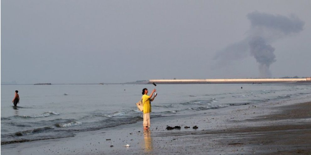 Smoke rises over the Strait of Hormuz near Bandar Abbas, Iran, as a woman stands along the shoreline following an explosion on July 13, 2026.
